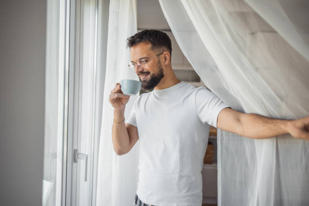 A young man enjoys his morning coffee while standing by the window and looking out into his yard