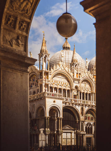 St. Mark’s Basilica in Venice, viewed through a stone arch. The background showcases the building’s rich architecture with its characteristic domes, and a crowd of tourists gathers below on the square