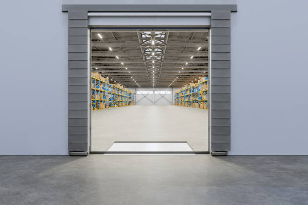 Outside View Of Distribution Warehouse With Open Roller Door. Cardboard Boxes On The Racks