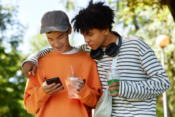 Black Boy Watching Male Asian Friend Using Phone while Walking in Park