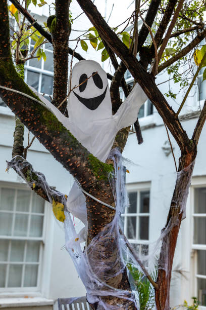 A charming white ghost decoration hangs from a tree in the front yard of a London house as part of a Halloween display. The playful ghost adds a lighthearted and whimsical touch to the festive scene, making it a perfect outdoor decoration for Halloween in October.