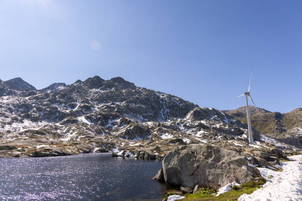 A wind turbine positioned on a snowy mountain slope near a calm lake, set against a clear sky.