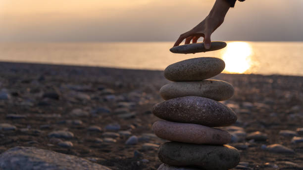 Close up woman's hand puts the last stone in the pyramid, copy space. Balanced pyramid of pebbles on the beach against the background of the sea at sunset.