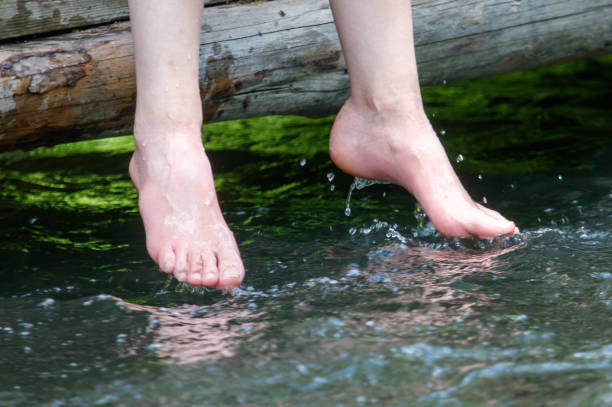 Bare feet touch the edge of a clear, flowing natural stream with a sandy and pebbled bank. The image captures a moment of peace and direct connection with nature.