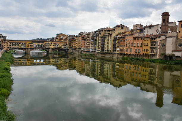 View of the Arno River in Florence, Tuscany, Italy, with a stone bridge and colorful historic buildings reflected in the calm water under a dramatic cloudy sky. Classic Italian cityscape.