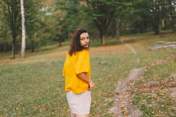 A woman in a bright yellow top walks through a park, smiling happily as she enjoys the beauty of nature and the warmth of the sun in autumn.