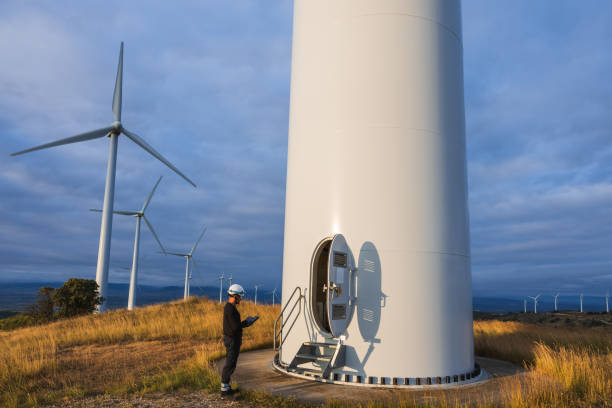 A wind turbine technician wearing a helmet and safety gear inspects a wind turbine using a digital tablet. the large wind turbines tower in the background against a clear sky, highlighting the importance of technology and maintenance in renewable energy.