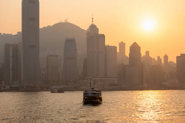 The setting sun appears behind various skyscrapers in Hong Kong, all dwarfed by the magnificent IFC building. In the foreground one of Hong Kong's iconic ferries has departed from the terminal at Tsim Sha Tsui and is making its way across the busy waters of Victoria Harbour.