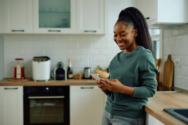 istock Happy black woman text messaging on cell phone in the kitchen. 2173119007