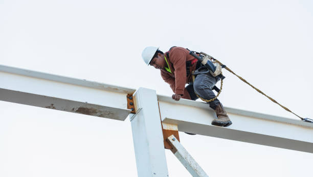 An ironworker on top of a steel building frame. The young Hispanic man is climbing on a roof beam wearing a hardhat, attached to a safety harness.