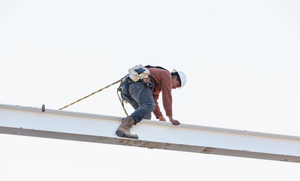 An ironworker on top of a steel building frame. The young Hispanic man is climbing on a roof beam wearing a hardhat, attached to a safety harness.
