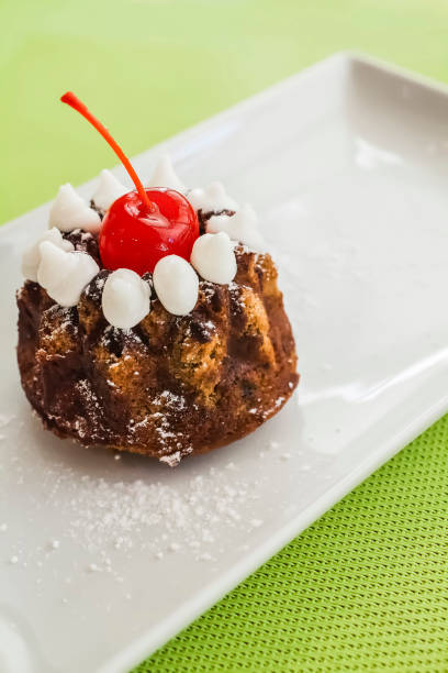 A beautifully presented mini bundt cake decorated with a bright red cherry on top, surrounded by dollops of white whipped cream, dusted with powdered sugar, and served on a white rectangular plate. The dessert is placed on a green textured background, highlighting its gourmet and appetizing appearance.