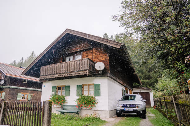 Bavaria, Germany - September 14, 2024 - A cozy Bavarian-style house with green shutters, a wooden balcony, a car parked in the driveway, and light rain falling in a rustic village.