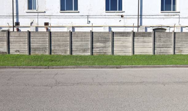 istock Concrete precast compound wall with barbed wire on top. White building on behind, strip of grass and road in front 2173275077