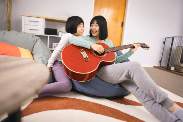 The daughter leans close to her mother, listening as she plays guitar while they relax together on a comfortable bean bag in a warm living room.