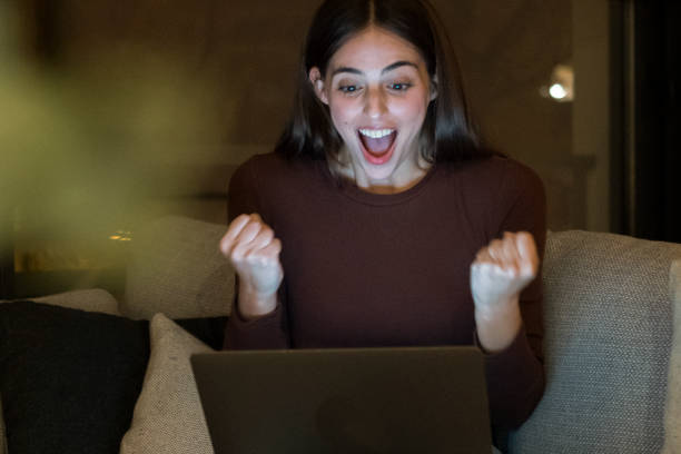 istock Excited young woman with a surprised expression looks at her laptop screen while sitting alone on a sofa at night. She is thrilled and happy, browsing the internet at home, celebrating a personal achievement 2173288891