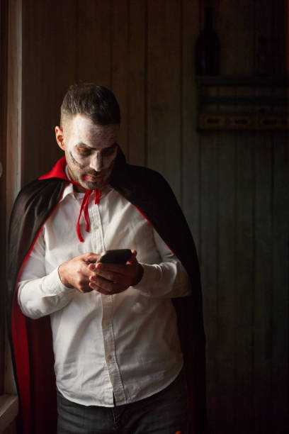 Carefree man in count Dracula costume text messaging on smart phone during Halloween at home. Copy space.
