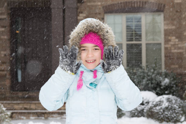 A cheerful young girl dressed warmly in winter clothing enjoys the snowfall in front of a brick home. Winter holidays concept.