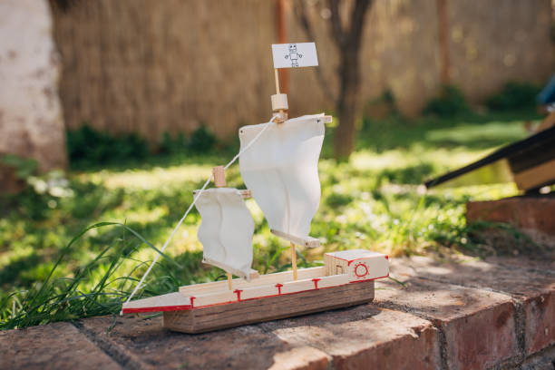 A detailed close-up of a handmade model sailboat with two sails, placed on a brick surface in an outdoor setting with grass and blurred background.