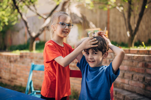A young girl enthusiastically places a wooden toy on a boy's head, captured in a delightful moment of shared laughter set in a vibrant garden.