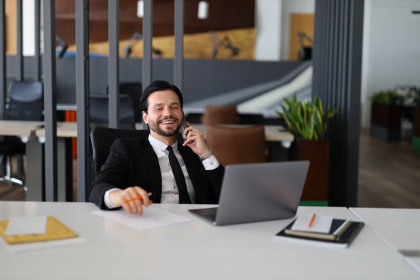 istock A man in a suit is talking on his cell phone while sitting at a desk. He is smiling and he is in a good mood 2173375876