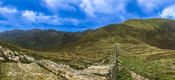 A scenic view from Slieve Donard in the Mourne Mountains, where the stone Mourne Wall stretches across the rugged terrain. The scene is framed by rolling hills and an expansive sky, creating a dramatic landscape.