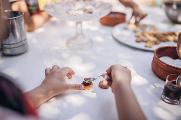 Young child carefully spreads homemade jam onto cookies in an sunny outdoor setting. The table has wooden bowls, a glass plate with cookies, and sunlight causing shadows.