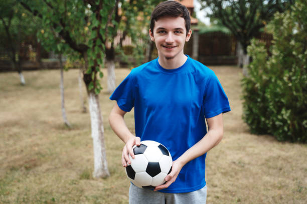 A young man in a blue t-shirt and gray shorts holds a soccer ball and smiles, with a grassy backyard and leafy trees in the background.