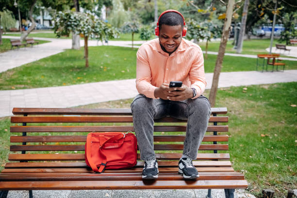 istock Young black man actively using mobile phone while resting on park bench in city park 2173399017