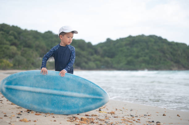 A 3 year old boy dressed in a long-sleeved swimsuit and white hat stands on a sandy beach, holding a blue surfboard with the ocean and mountainous landscape in the background. Tropical summer destination in Koh Samed Thailand