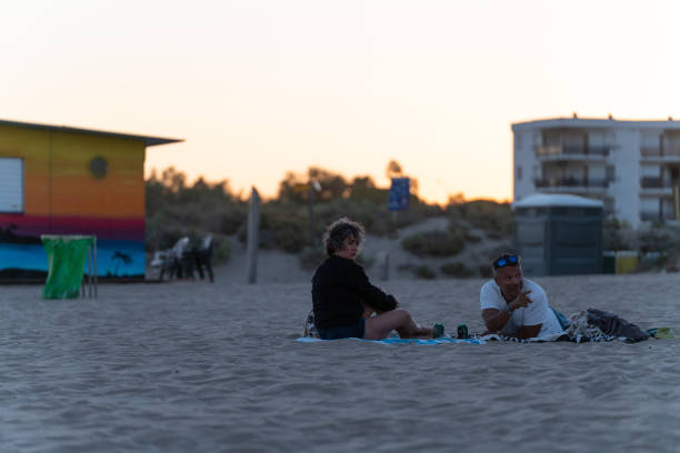 A man and a woman sit on a blanket on the beach during sunset, enjoying the calm atmosphere. The sky glows with soft pastel colors as the day fades. In the background, a colorful building and residential area add contrast to the peaceful beach setting.