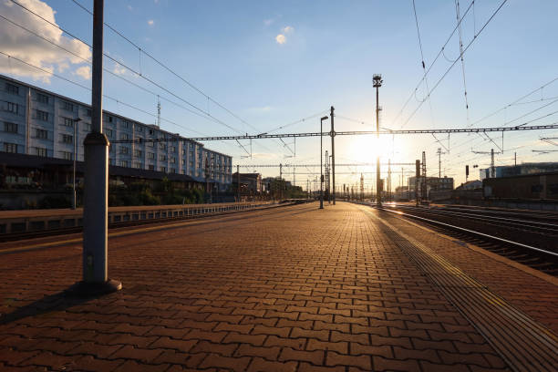 Prague Liben station platform at sunset, bathed in golden light. Overhead power lines stretch into the distance, casting long shadows across the brick-paved platform. The industrial backdrop and calm atmosphere evoke a serene, quiet moment.
