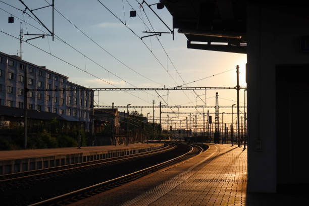 Prague Liben railway station at sunset, with tracks curving under a web of overhead power lines. The warm light casts long shadows across the platform, highlighting the station's industrial architecture.