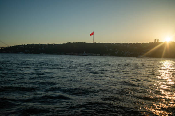 A breathtaking view of the Istanbul Bosphorus at sunset, featuring the iconic Turkish flag waving proudly. The golden light reflects on the water, highlighting the city's historic skyline and creating a serene, picturesque scene.