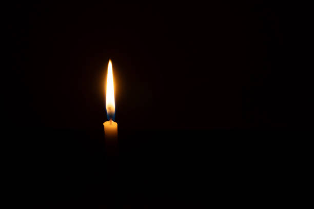 A single burning candle flame or light is glowing on a small orange candle on black or dark background on table in church for Christmas, funeral or memorial service with copy space