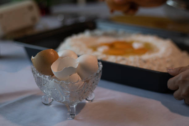 Photography with copy space of some fragile broken egg shells in a glass container next to a baking sheet with eggs and flour for the preparation of dough.