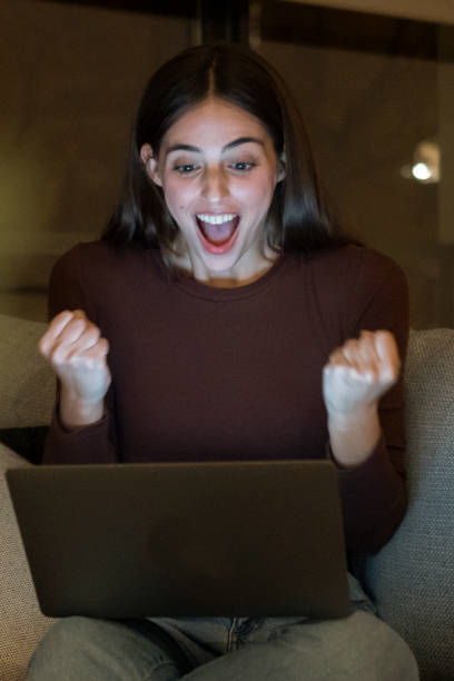 istock Excited young woman with a surprised expression looks at her laptop screen while sitting alone on a sofa at night. She is thrilled and happy, browsing the internet at home, celebrating a personal achievement 2173487638