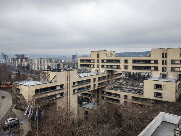 A modern architectural structure with interconnected blocks set against a cityscape with numerous high-rise buildings under an overcast sky.
