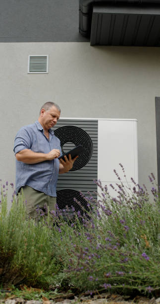 istock A homeowner examines an external heat pump unit using a tablet, standing amid lavender plants. 2173505263