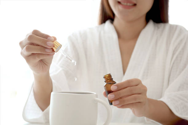 Asian woman in white bathrobe dripping naturopathic medicine on her drink glass. Homeopathic capacity to heal itself, naturopathy and alternative medicine Asian woman in white bathrobe dripping naturopathic medicine on her drink glass. Homeopathic capacity to heal itself, naturopathy and alternative medicine Caffeine Serum for Eyes stock pictures, royalty-free photos & images