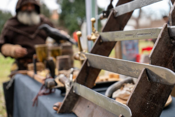 Several medieval swords displayed on a wooden stand at a theme fair. Sword blades decorated with detailed engravings. In the background, a man dressed in medieval clothing observing the exhibition. The scene captures the essence of a medieval fair, with a focus on tradition and warrior history.