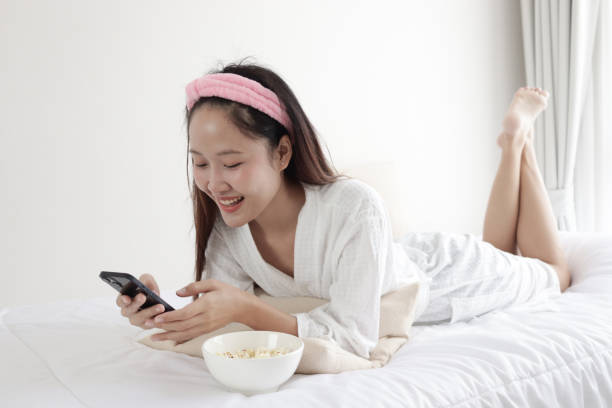 istock Happy Asian woman in white bathrobe using smartphone and eating popcorn snack while lying on bed at home. Relaxing beautiful girl using her digital device mobile phone for entertainment on bed. Young female bed rotting on weekend. 2173522500