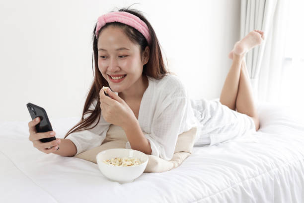 istock Happy Asian woman in white bathrobe using smartphone and eating popcorn snack while lying on bed at home. Relaxing beautiful girl using her digital device mobile phone for entertainment on bed. Young female bed rotting on weekend. 2173522507