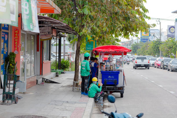 Construction workers are taking a break at street food market stall in street Wanghin Road in Bangkok Ladprao. Market stall is a pushcart tricycle and is selling iced drinks. A group of construction workers is standing  at market stall or sitting down on sidewalk. On street is some traffic. In background are trees along street