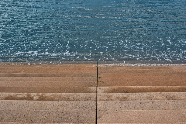 A view of concrete steps leading down to a calm body of water. The steps are dry and clean, with gentle waves lapping at the base. The water is clear and reflects the light, creating a serene atmosphere.