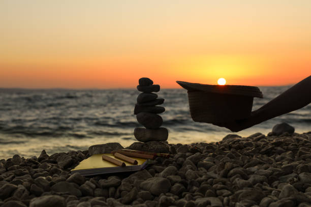 A woman's hand holds a straw hat at sunset, into which the sun is setting, and which lies next to a cairn of stones, a notebook and pencils.