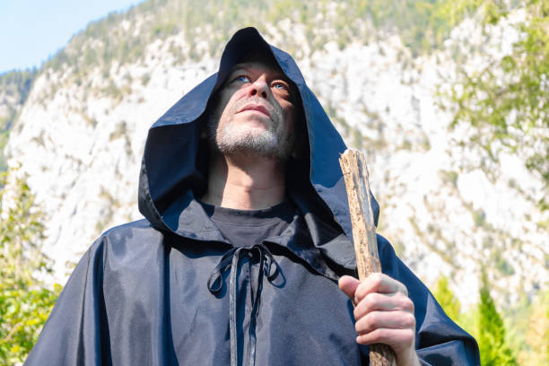 istock A monk in a dark cloak stands with a staff looking pensively into the sky against a backdrop of towering mountains on a sunny day surrounded by nature. 2173606473