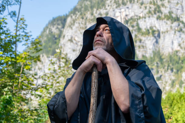 istock A monk in a dark cloak stands with a staff looking pensively into the sky against a backdrop of towering mountains on a sunny day surrounded by nature. 2173606541