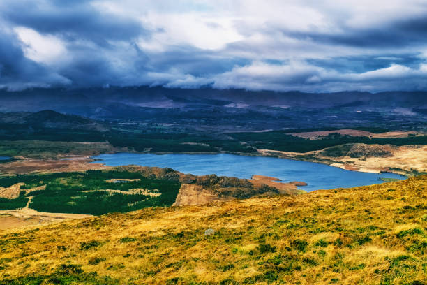 A panoramic view of a lake nestled between hills, with a blue sky and a dramatic mountain range in the distance, covered in clouds.