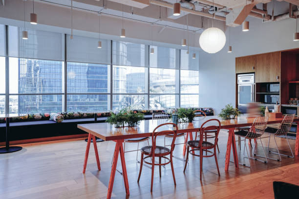 A vibrant and modern co-working space with a mix of comfortable lounge seating and work tables, offering a stylish yet functional environment for collaboration and productivity. The space features a large open area with a combination of wooden furniture, colorful cushions, and indoor plants that create a welcoming atmosphere. The industrial ceiling design with exposed pipes adds a contemporary touch, while the large windows allow ample natural light to flood the room. The blend of traditional and modern elements, such as the wooden partition screens and sleek furniture, contributes to a dynamic and creative workspace.